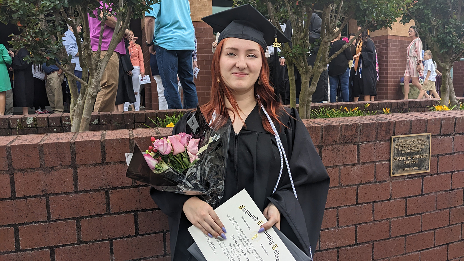 A graduating student stands at the Cole Auditorium holding her diploma and a boquet of flowers.